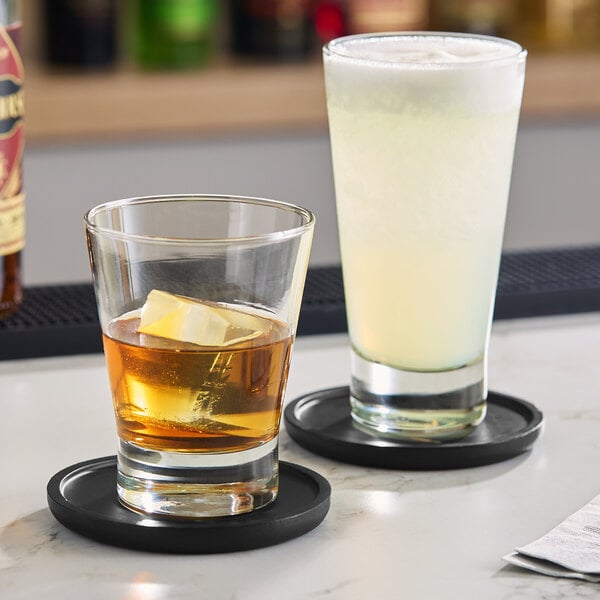 A clear Acopa Fusion 12 oz. rocks glass filled with a brown beverage and ice, placed on a black coaster.