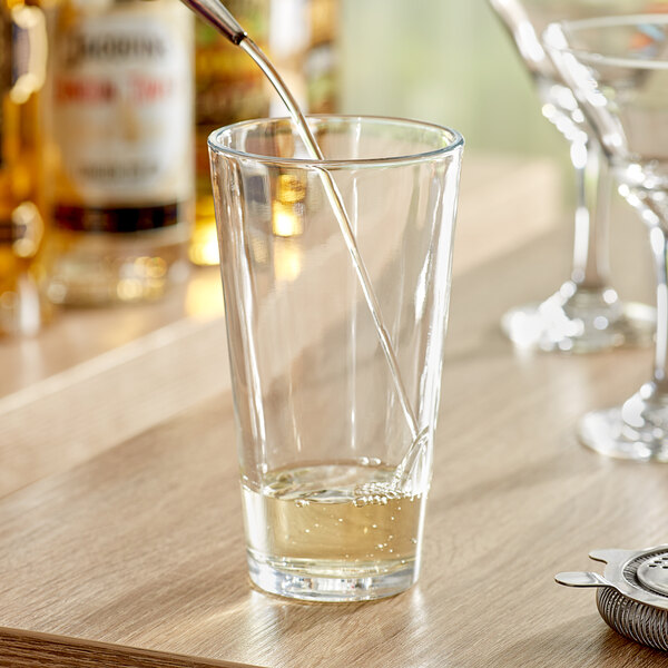 A clear 20 oz. mixing glass partially filled with liquid, shown on a wooden surface with bar tools and bottles in the background.