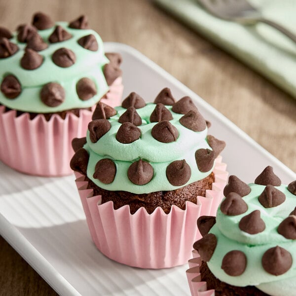 A chocolate cupcake with light green peppermint frosting topped with dark chocolate chips, presented in a pink cupcake liner.