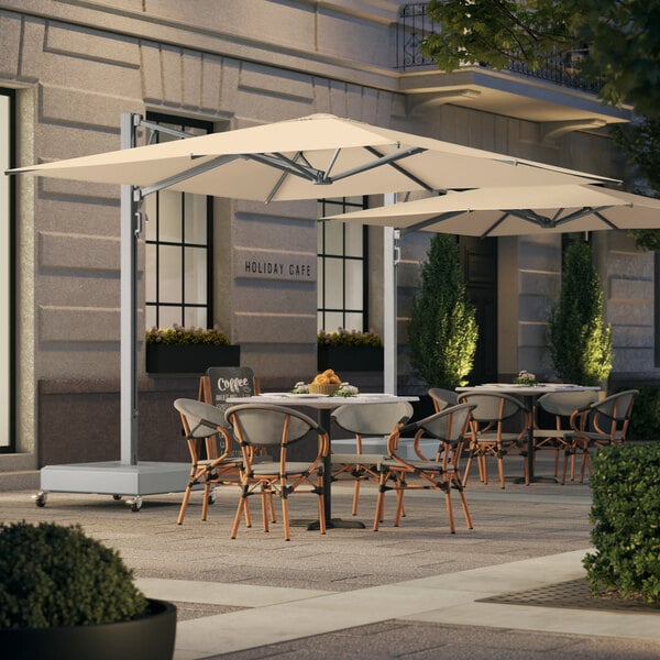 A large square sand-colored cantilever umbrella with a silver aluminum frame and hydraulic lift, positioned over outdoor café tables and chairs.