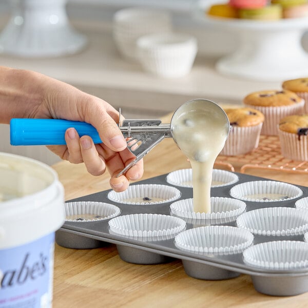A hand uses a blue-handled scoop to portion muffin batter into paper-lined muffin tins.