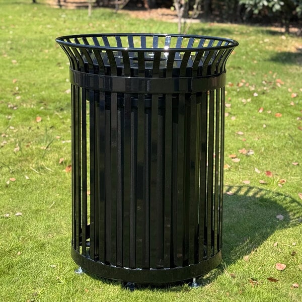 A black, round, slatted metal outdoor trash can is placed on a grassy area.