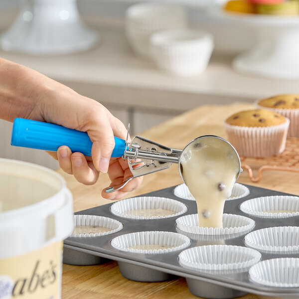 A hand is using a blue-handled scoop to portion muffin batter with chocolate chips into paper-lined muffin tins.
