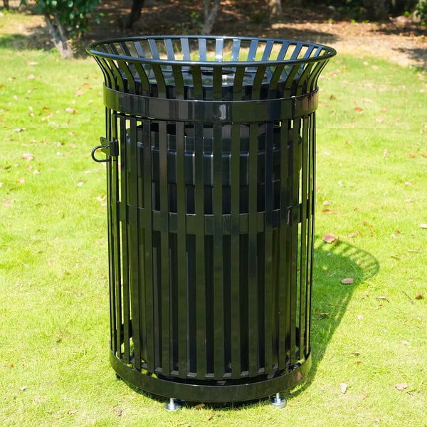 A black, round, slatted metal outdoor trash can is standing on grass in a park-like setting.