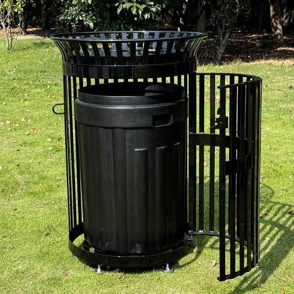 A large black outdoor trash can with a slatted steel enclosure, recycling lid, and an open door, placed on grass in a park setting.
