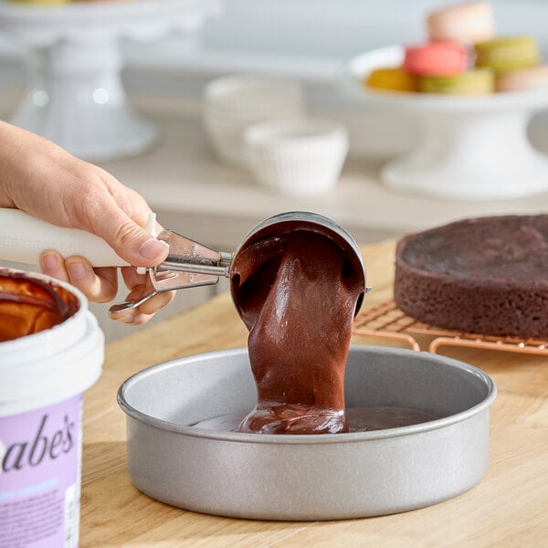 A hand is scooping chocolate cake batter into a round baking pan.
