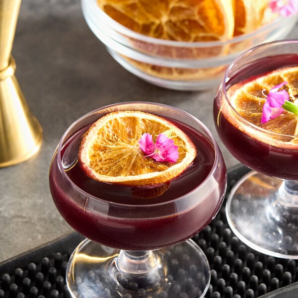 A glass of dark red cocktail garnished with a dried orange slice and a small pink flower.