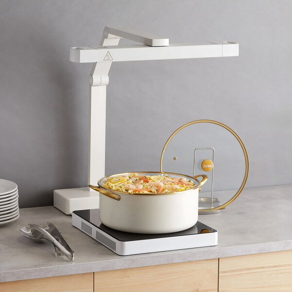 A white and gold accent stainless steel insert pan filled with food, placed on an induction cooktop with a glass lid nearby.