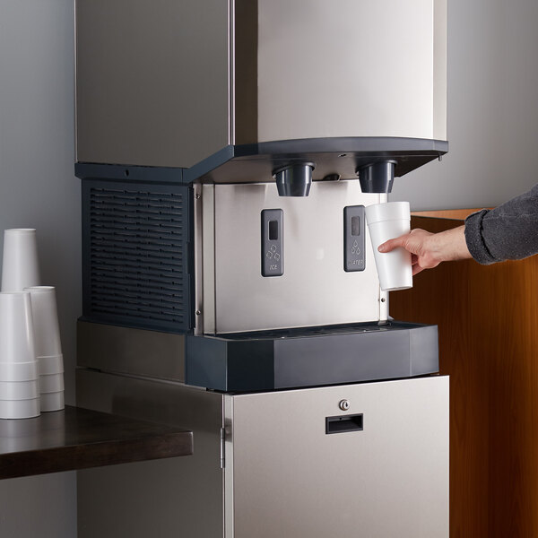 A stainless steel countertop nugget ice machine and water dispenser with hands-free dispensing, shown in use with a person holding a cup under the dispenser.