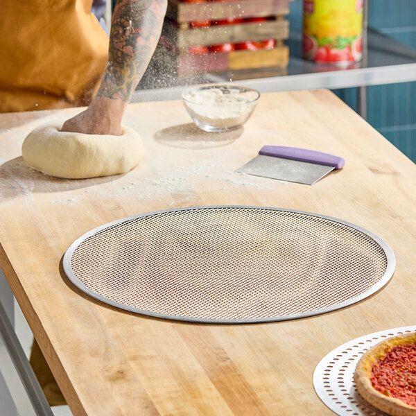 A round, heavy duty expanded aluminum pizza screen on a wooden countertop.