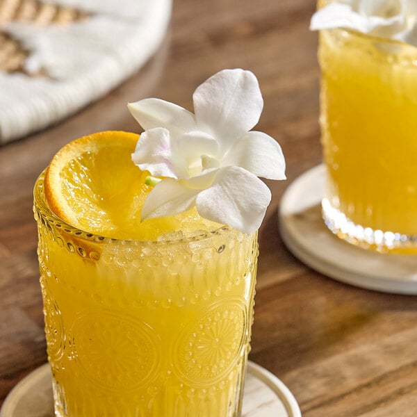 A glass of orange beverage garnished with a white edible orchid and an orange slice.