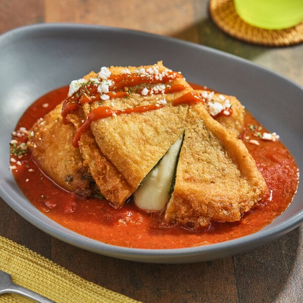 A plate of breaded and fried chile relleno stuffed with cheese, served with red sauce and garnished with a drizzle of sauce and crumbled cheese.