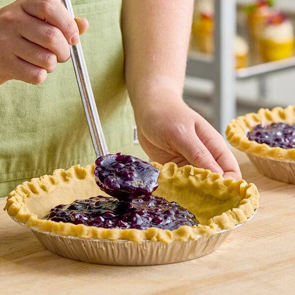A pie crust being filled with blueberry filling from a metal ladle.