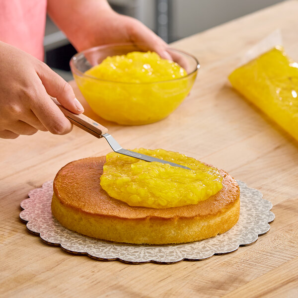 A person spreading yellow pineapple pastry filling onto a round cake layer with a spatula.