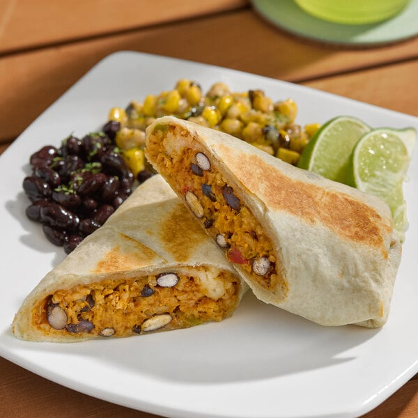A chicken fajita burrito cut in half, served with black beans, corn, and lime wedges on a white plate.