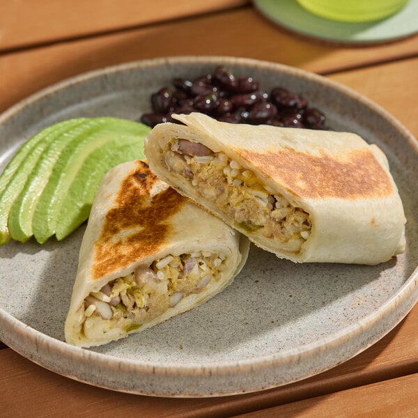 A plate with a pork carnitas burrito cut in half, served with sliced avocado and black beans.