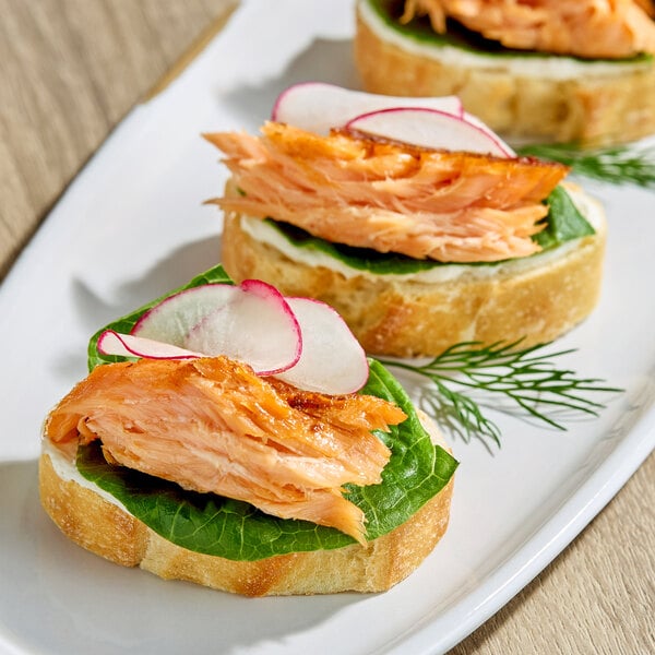 An appetizer featuring slices of bread topped with leafy greens, honey smoked salmon fillet, and thinly sliced radishes, served on a white plate.