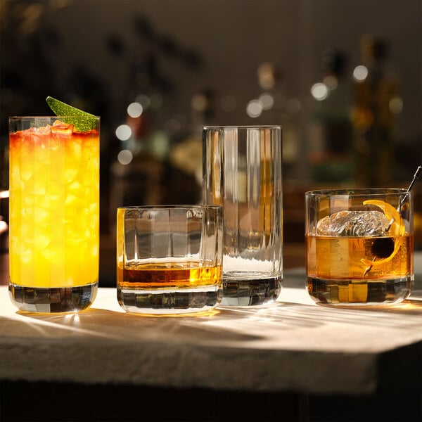 A set of clear glass beverage glasses, including both tall and short styles, displayed on a bar surface with various drinks inside.