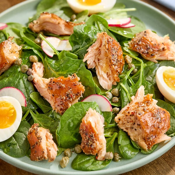 A plate of salad topped with cracked pepper honey smoked salmon pieces, spinach leaves, capers, radish slices, and halved boiled eggs.