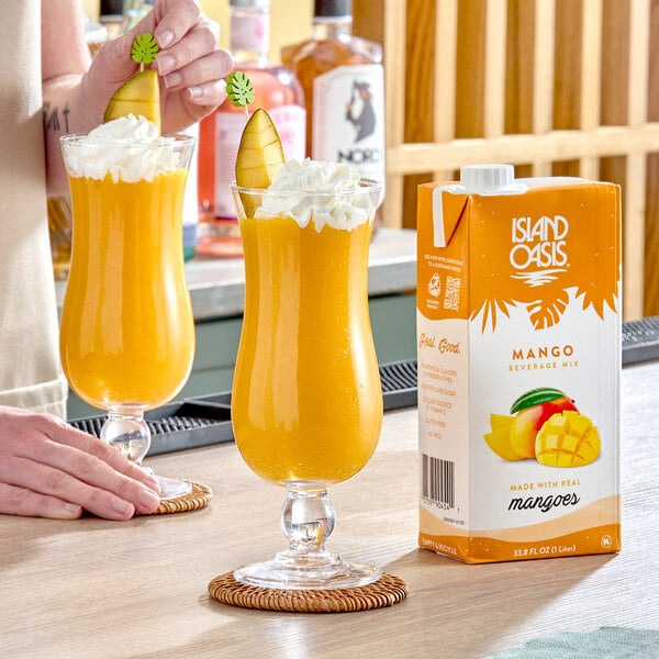 A carton of Island Oasis Mango Beverage Mix next to two glasses of mango smoothie topped with whipped cream and garnished with mango slices.