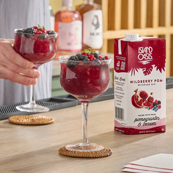A 1-liter carton of Island Oasis Wildberry Pom Beverage Mix is shown next to a glass of red frozen drink garnished with fresh berries.