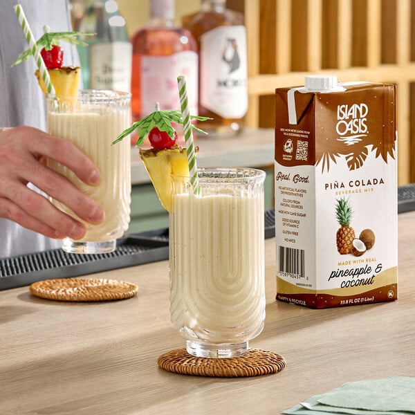 A 1-liter carton of Island Oasis Pina Colada Beverage Mix is shown next to two glasses of prepared pina colada garnished with pineapple and cherry.