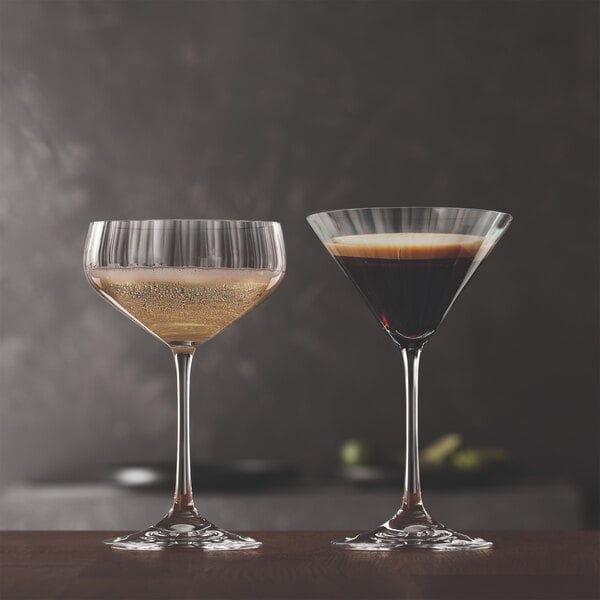 A pair of Spiegelau Lifestyle 7.5 oz. martini glasses, one filled with a light sparkling beverage and the other with a dark cocktail, displayed on a wooden surface.