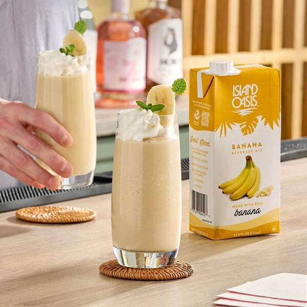 A 1-liter carton of Island Oasis Banana Beverage Mix next to two glasses of banana smoothie garnished with whipped cream, banana slices, and mint.