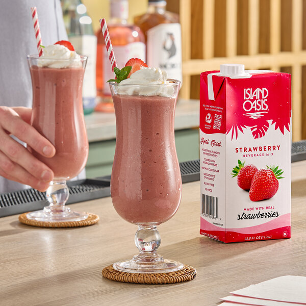 A carton of Island Oasis Strawberry Beverage Mix next to two glasses of strawberry smoothies topped with whipped cream and garnished with strawberries.