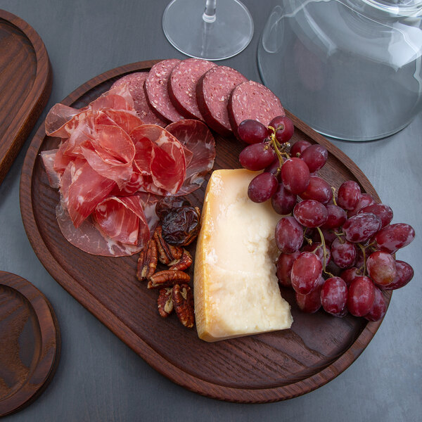 A dark brown oval ash wood serving board displaying cheese, grapes, cured meats, nuts, and dried fruit.