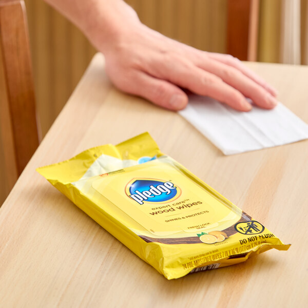 A package of SC Johnson Pledge Expert Care lemon wood wipes on a wooden surface with a hand using a wipe nearby.