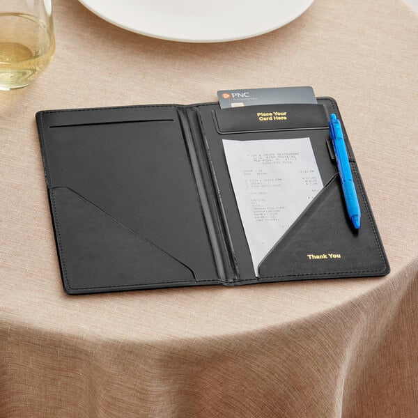 A black vinyl guest check presenter with pockets for a receipt, pen, and card, displayed on a table.