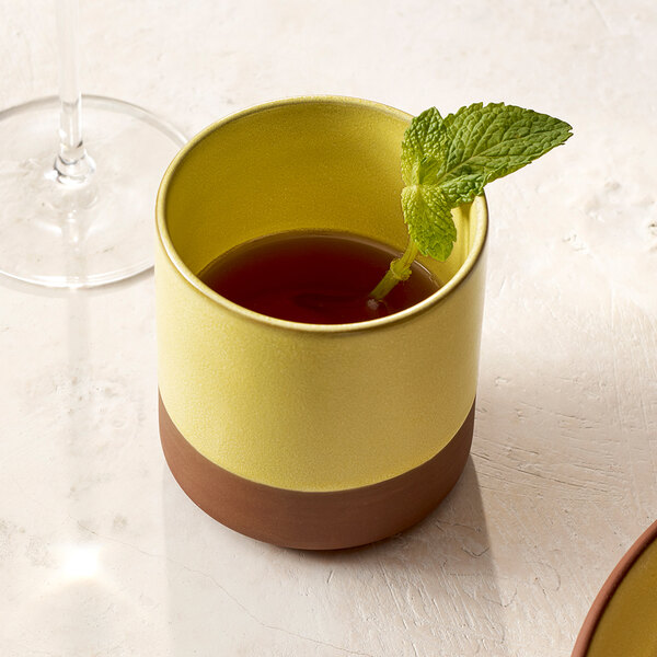 A round saffron-colored stoneware cup with a brown base, filled with a dark beverage and garnished with a mint sprig.