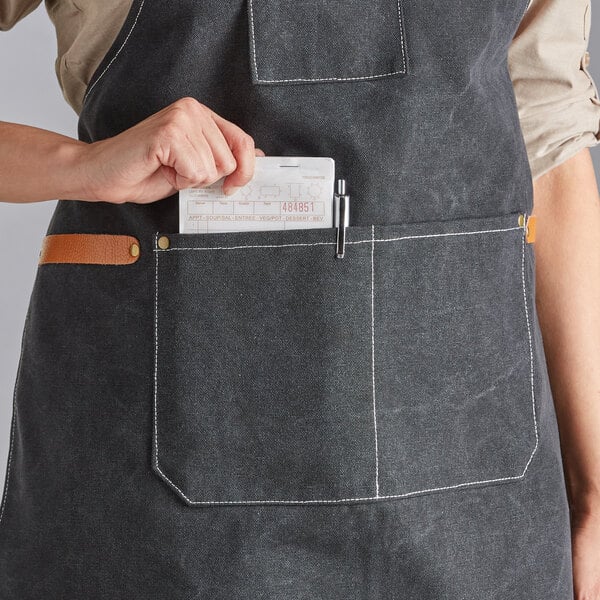 A black heavy weight canvas cross-back bib apron with three pockets, shown being worn by a person placing an order pad and pen in the front pocket.