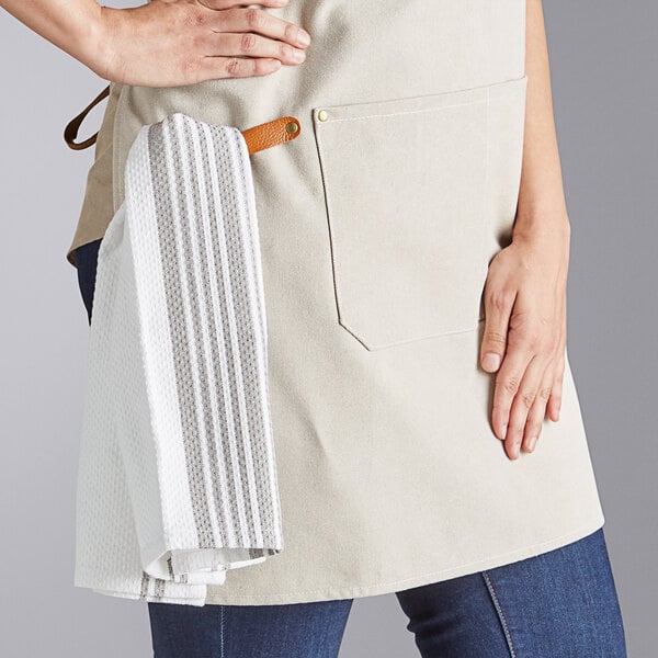 A cream-colored heavy weight canvas cross-back bib apron with three pockets, shown with a towel hanging from a leather loop.