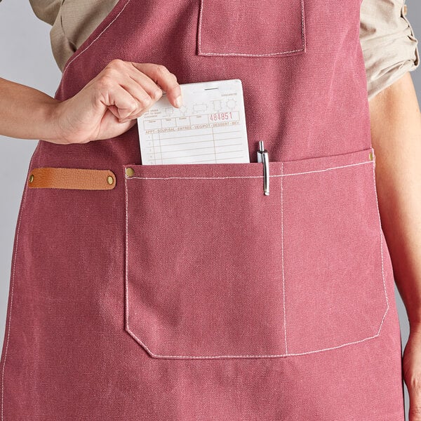 A wine-colored heavy weight canvas cross-back bib apron with three pockets, shown being worn with a notepad and pen in the front pocket.
