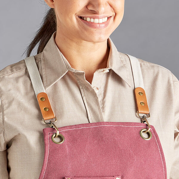 A person wearing a wine-colored heavy weight canvas cross-back bib apron with leather and fabric straps and metal grommets.