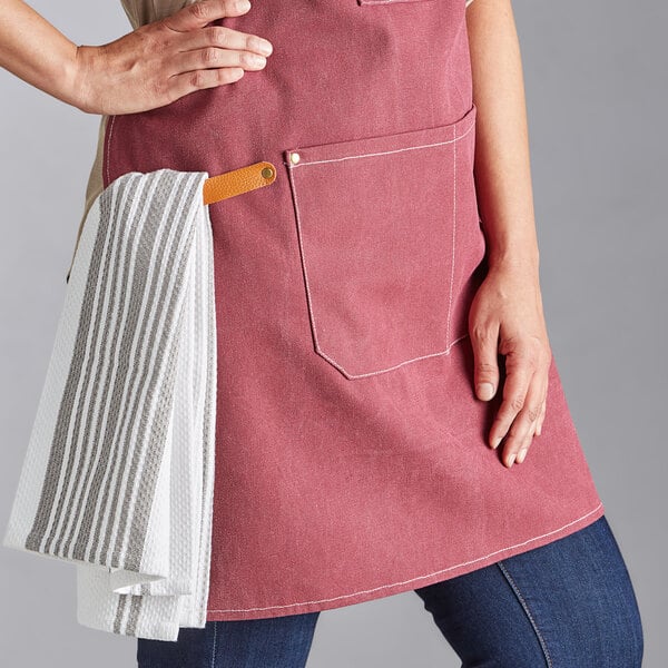 A person wearing a wine-colored canvas cross-back bib apron with three pockets and a striped towel hanging from a leather loop.