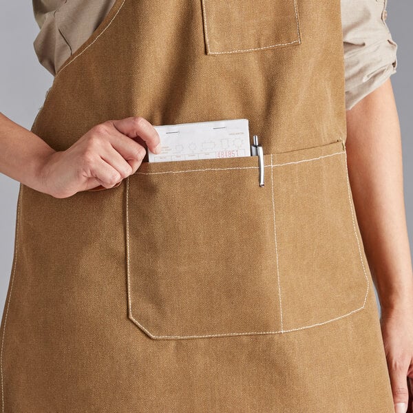 A person wearing a mocha-colored heavy weight canvas cross-back bib apron with three pockets, placing a notepad and pen in the front pocket.