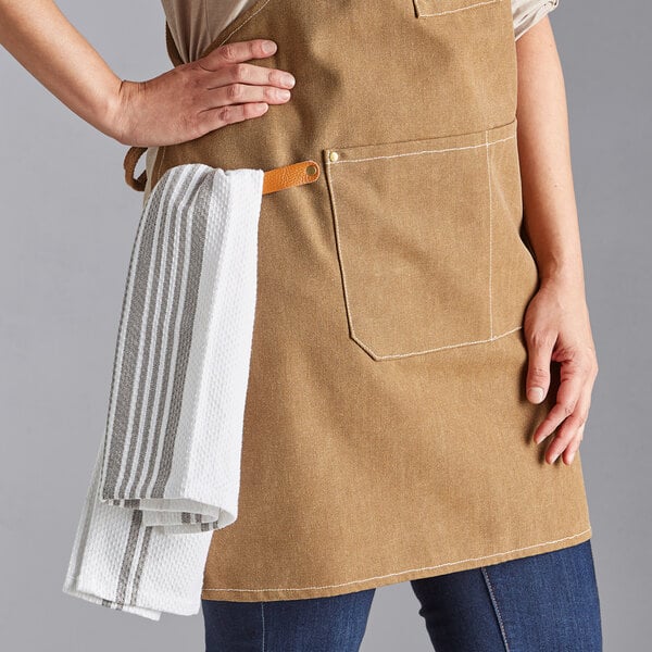 A mocha-colored heavy weight canvas cross-back bib apron with three pockets, shown with a towel hanging from a side loop.