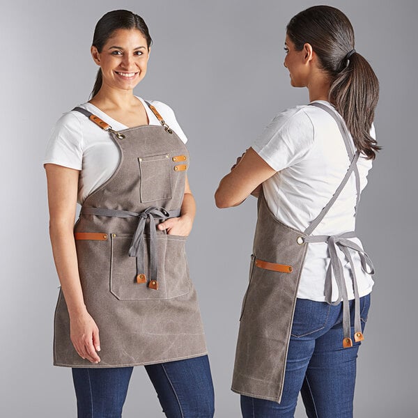 A heavy weight canvas cross-back bib apron in pebble color with three pockets, shown from the front and back on a model.