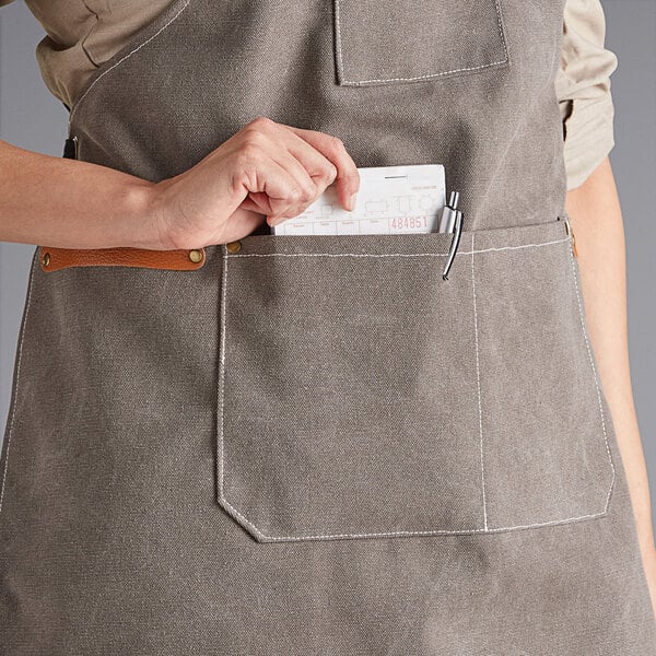 A person wearing a gray canvas cross-back bib apron with three pockets, placing an item into the front pocket.