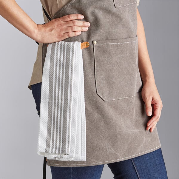 An individual wearing a gray canvas cross-back bib apron with three pockets and a white towel attached to the side.