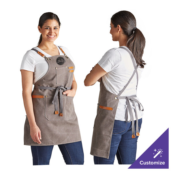 A heavy weight canvas cross-back bib apron in pebble color with three pockets, shown from the front and back on a model.