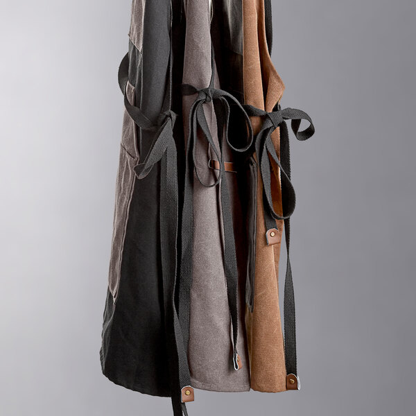 A pair of heavy-duty canvas grilling aprons with utility pockets in charcoal, gray, and brown colors hanging side by side.