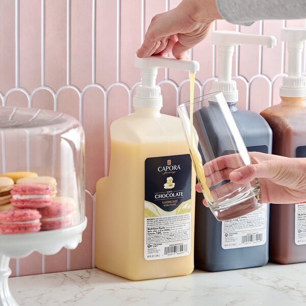 A large bottle of Capora white chocolate flavoring sauce with a pump dispenser being used to pour sauce into a glass.