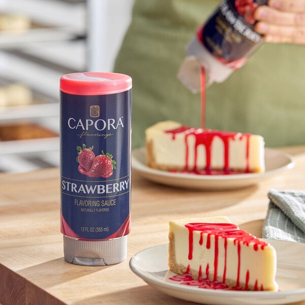 A 12 fl. oz. bottle of Capora Strawberry Flavoring Sauce placed on a wooden surface next to cheesecake slices drizzled with the sauce.