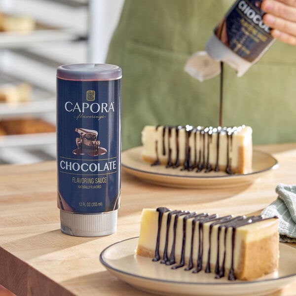 A 12 fl. oz. bottle of Capora Chocolate Flavoring Sauce displayed on a table with cheesecake slices drizzled with chocolate sauce in the background.