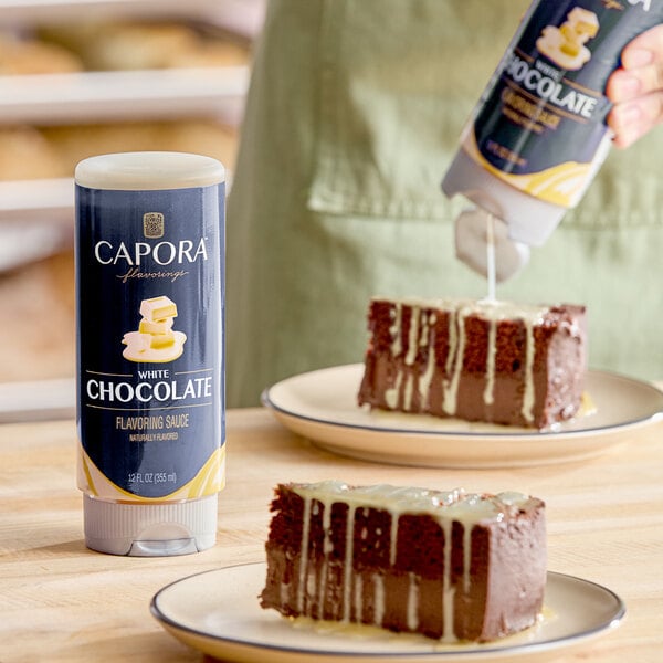 A bottle of Capora White Chocolate Flavoring Sauce next to slices of chocolate cake being drizzled with the sauce.