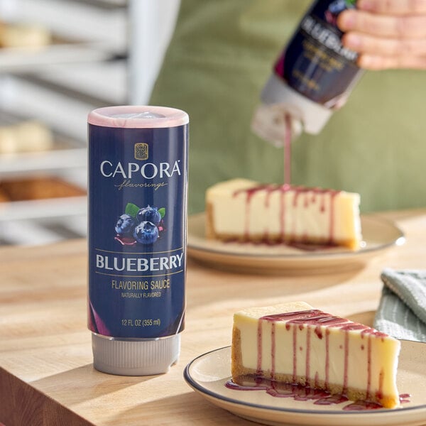 A 12 fl. oz. bottle of Capora Blueberry Flavoring Sauce, shown being used to drizzle sauce over slices of cheesecake.
